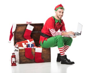 gülümseyen adam ahşap kutu üzerinde oturan ve üzerinde beyaz izole dizüstü kullanarak Noel elf kostümü 