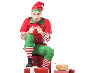 heyecanlı adam hediyeleri yığını üzerinde oturan ve video üzerinde beyaz izole oyun oynarken Noel elf kostümü