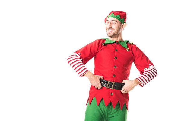 cheerful man in christmas elf costume looking at camera with hands on hips isolated on white