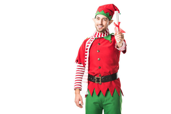 smiling man in christmas elf costume holding wishlist roll and looking at camera isolated on white