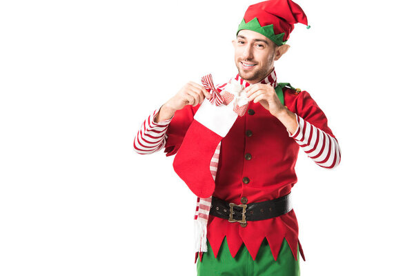 smiling man in christmas elf costume putting present in red christmas stocking and looking at camera isolated on white