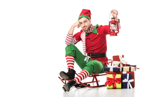 man in christmas elf costume sitting on sleigh near pile of presents and holding red lantern isolated on white