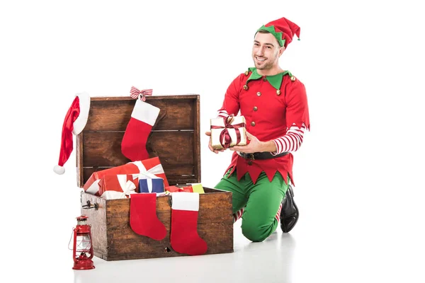 クリスマス エルフ衣装プレゼントの木製箱のそばに座って ギフト白で隔離で笑みを浮かべて男 — ストック写真