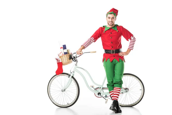 エルフ衣装がいっぱい入ったかごが付いているバイクの近く股関節立って手にプレゼントに孤立したホワイト クリスマスで幸せな笑みを浮かべて男 — ストック写真