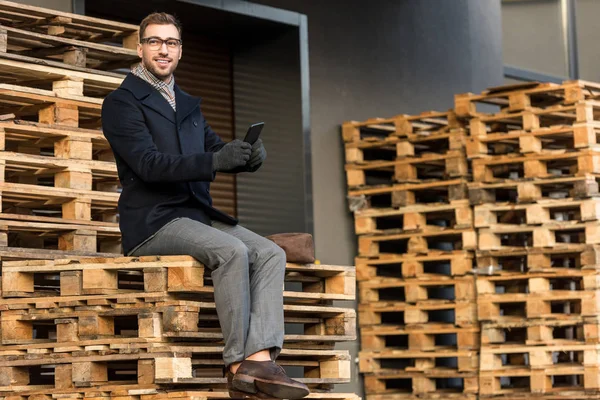 Handsome smiling man using smartphone and sitting on wooden pallets — Stock Image Handsome Smiling Man Using Smartphone Sitting Wooden Pallets Stock Picture