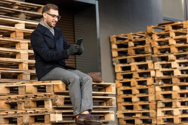 Handsome smiling man using smartphone and sitting on wooden pallets — Stock Image Handsome Smiling Man Using Smartphone Sitting Wooden Pallets Royalty Free Stock Photos