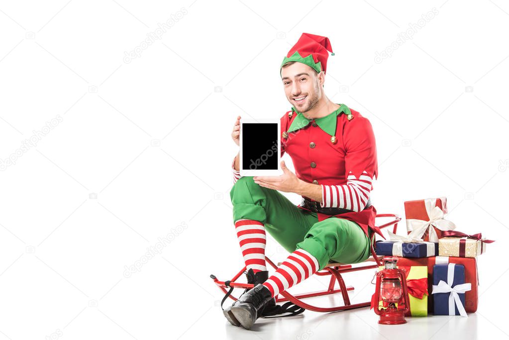 man in christmas elf costume sitting on sleigh and holding tablet with blank screen isolated on white
