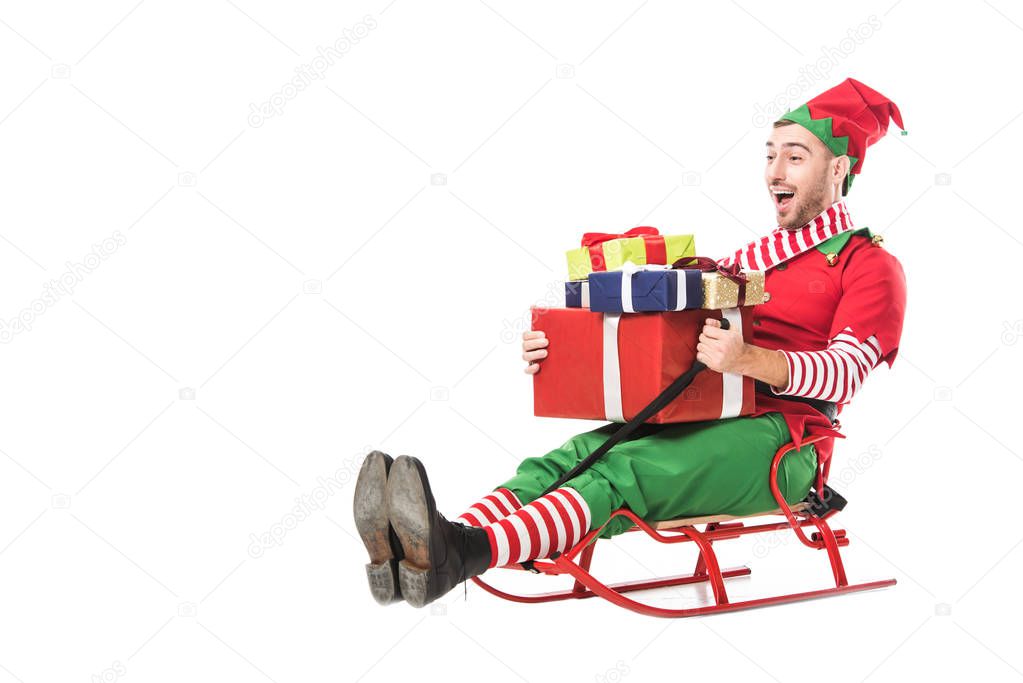 cheerful excited man in christmas elf costume holding pile of presents and riding sleigh isolated on white