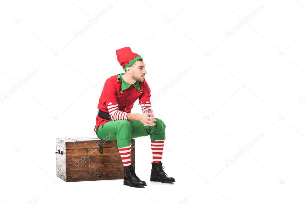 pensive man in christmas elf costume sitting on wooden chest isolated on white