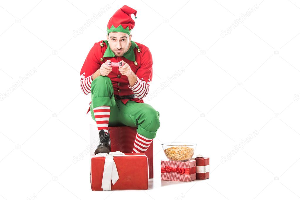 man in christmas elf costume sitting on pile of presents and playing video game isolated on white