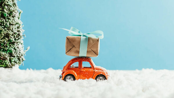 side view of toy red car with christmas gift box riding by snow made of cotton on blue background