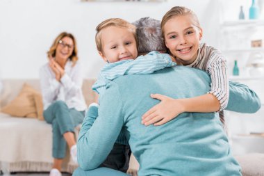 selective focus of smiling kids looking at camera and hugging grandfather at home clipart