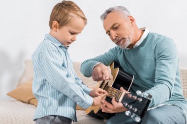 man pointing by finger while teaching grandson playing on acoustic guitar at home clipart