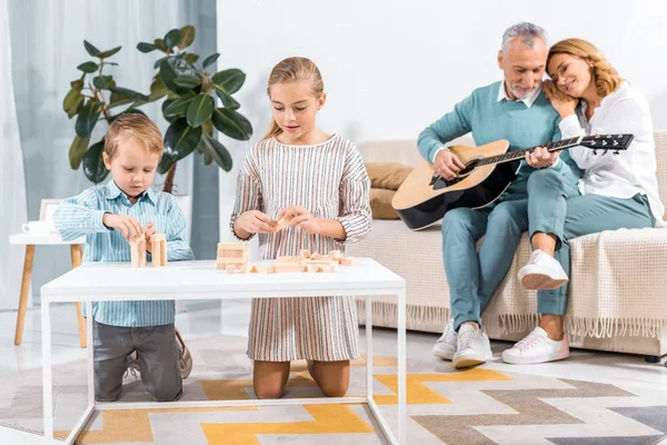 祖父母の家で後ろにギターで遊んでいる間ブロック木製ゲーム タワーを遊んでいる子供たちの選択と集中 — ストック写真