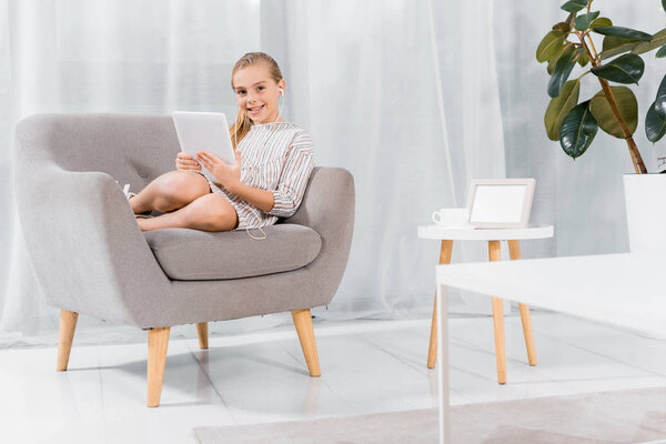 cheerful little child in earphones sitting on armchair with digital tablet at home