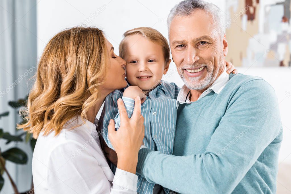 selective focus of cheerful middle aged couple holding adorable grandson and kissing him in cheeks