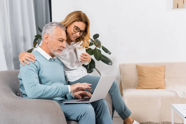 中间男人使用笔记本电脑的侧视图 而他微笑的妻子坐在附近与咖啡在家里 — 图库照片