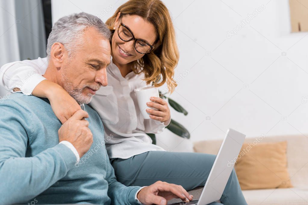 cheerful middle aged man using laptop while his smiling wife sitting near with coffee at home