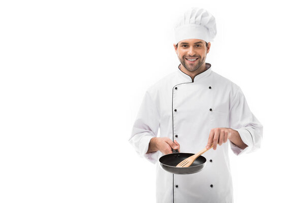 smiling young chef holding frying pan and wooden spatula isolated on white