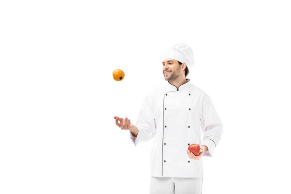 smiling young chef juggling with tomato and bell pepper isolated on white