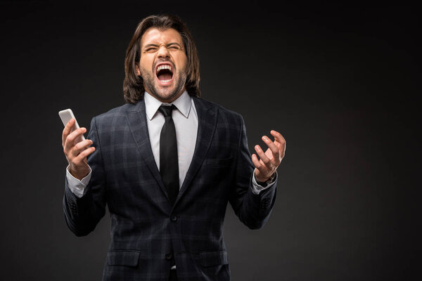 разъяренный молодой бизнесмен держит смартфон и кричит изолированно на черном
