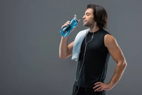 微笑的肌肉发达的人用毛巾喝水从运动瓶隔离在灰色 — 图库照片