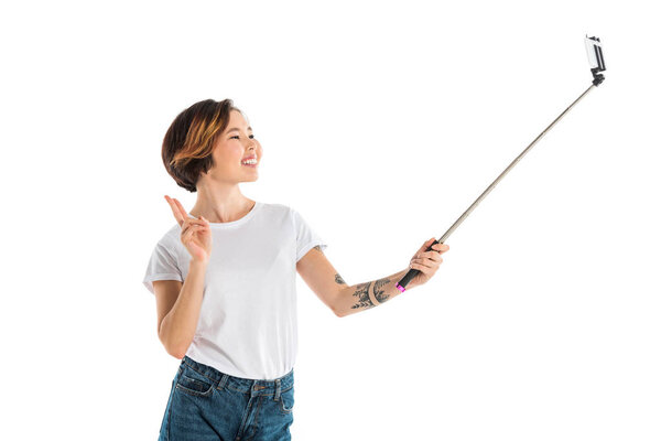 attractive smiling young woman taking selfie on smartphone using selfie stick isolated on white