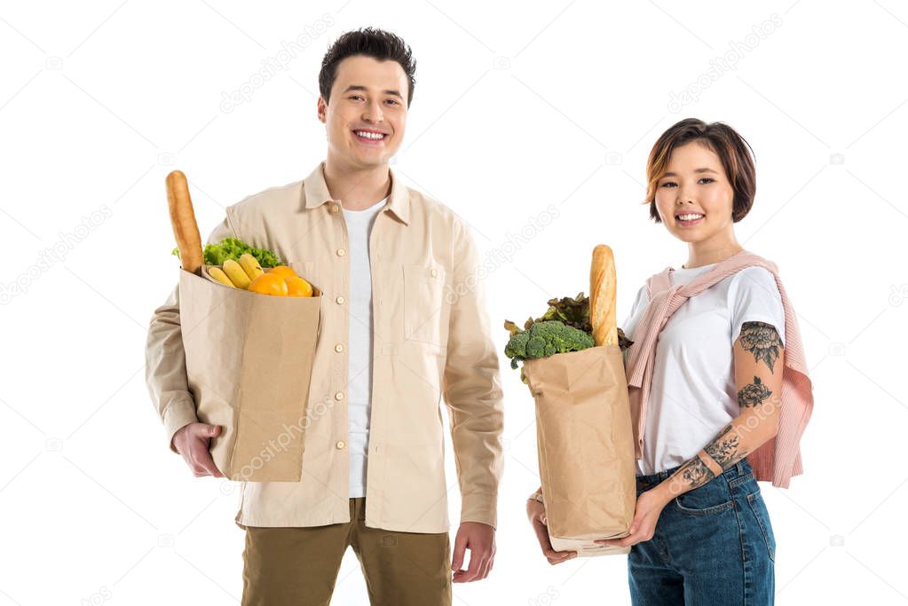 happy husband and wife holding grocery bags and looking at camera isolated on white