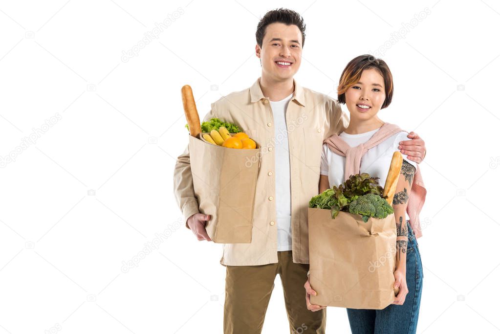 smiling husband and wife holding grocery bags and looking at camera isolated on white