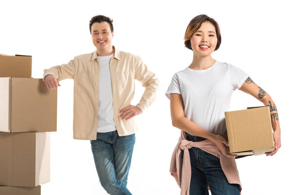 smiling couple looking at camera and holding cardboard boxes isolated on white, moving to new house concept