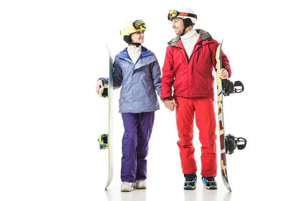 smiling couple in ski suits holding snowboards and looking at each other isolated on white