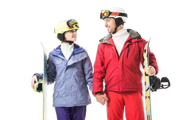 Couple in ski clothes with snowboards holding hands,  looking at each other and smiling isolated on white
