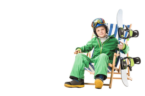 Preteen boy in green ski suit with snowboard sitting in deck chair isolated on white