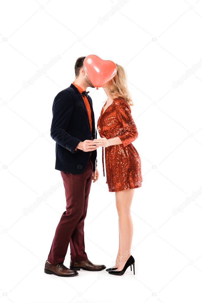 Attractive couple holding hands and kissing while hiding faces behind red balloon isolated on white