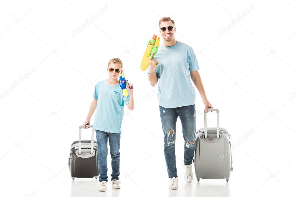 Father and son walking in sunglasses and holding water guns and luggage isolated on white