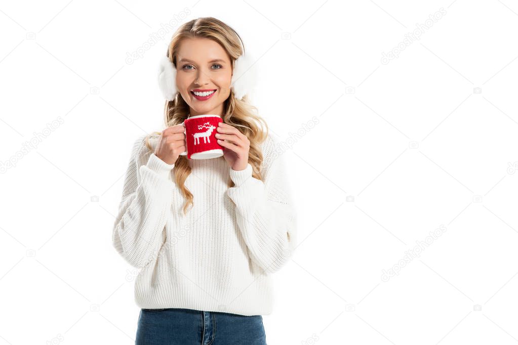 attractive smiling girl in winter earmuffs holding cup with hot coffee isolated on white