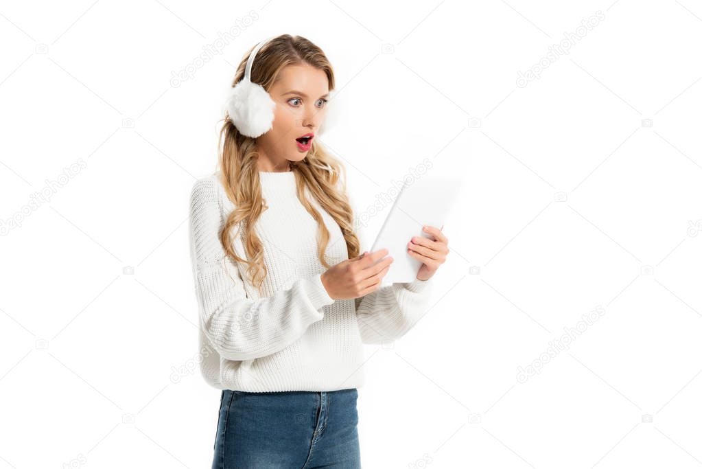 shocked blonde girl in stylish earmuffs looking at digital tablet isolated on white 