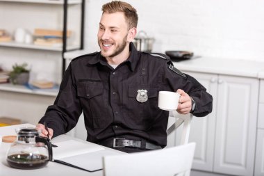 mutfak masasında oturan, not defterinde yazılı ve kahve içme polis gülümseyen