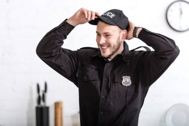 handsome police officer smiling and putting on cap at kitchen clipart