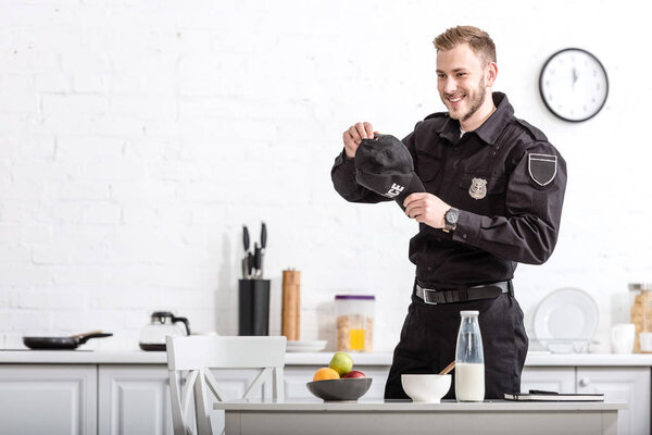 handsome police officer with cap in hands smiling at kitchen