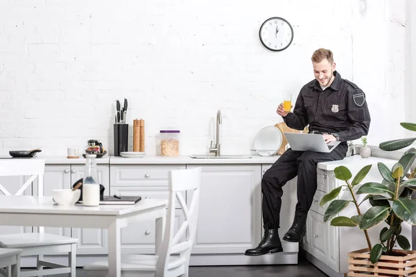年轻的警察坐在桌子上 喝橙汁 在厨房里使用笔记本电脑 — 图库照片