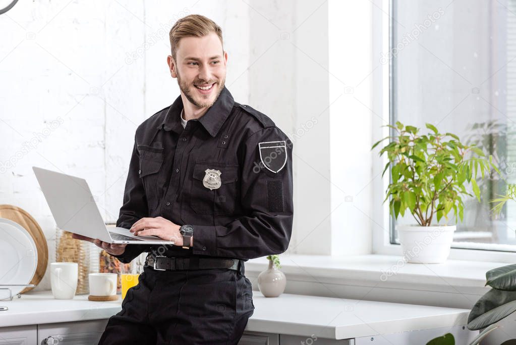 handsome policeman smiling and using laptop at kitchen