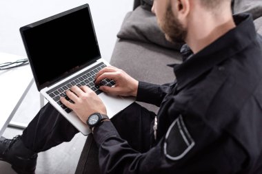 cropped view of police officer sitting on couch and typing on laptop keyboard clipart