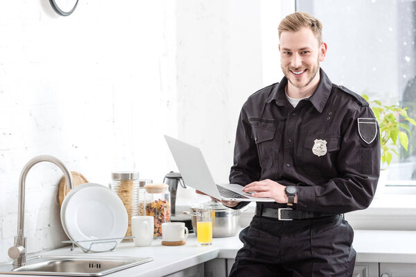 Smiling police officer standing with laptop at kitchen