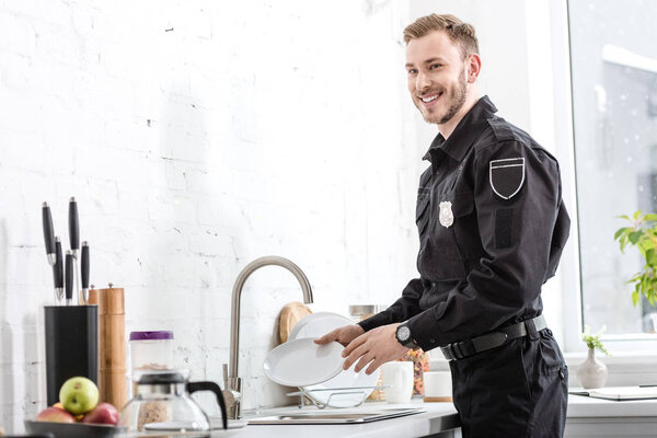 Handsome police officer smiling and washing plate at kitchen