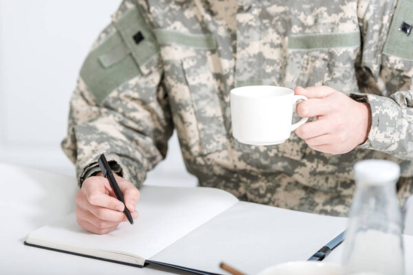 cropped view of man in military uniform drink coffee and writing in notebook