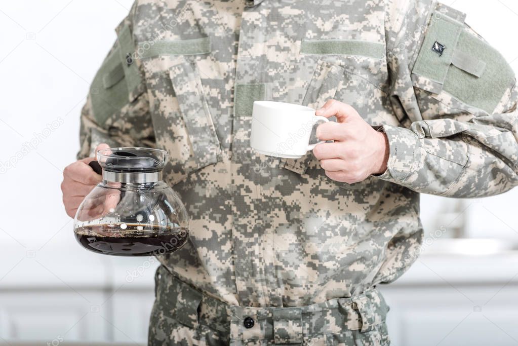 partial view of army soldier holding cup of coffee and pot in kitchen