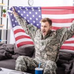 兴奋的陆军士兵坐在沙发上, 欢呼和自豪地拿着美国国旗