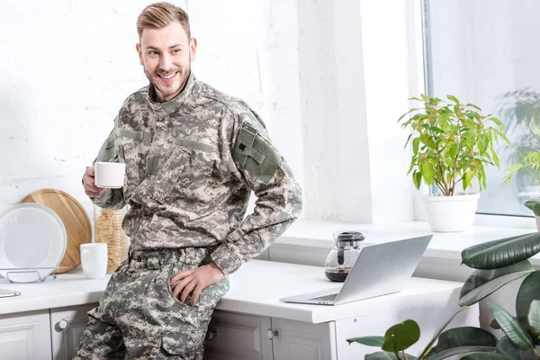 英俊的士兵拿着一杯热咖啡与笔记本电脑在厨房台面上 — 图库照片