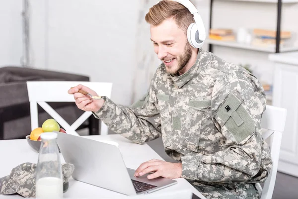微笑的士兵在耳机使用笔记本电脑在厨房的桌子上 而有早餐 — 免费的图库照片